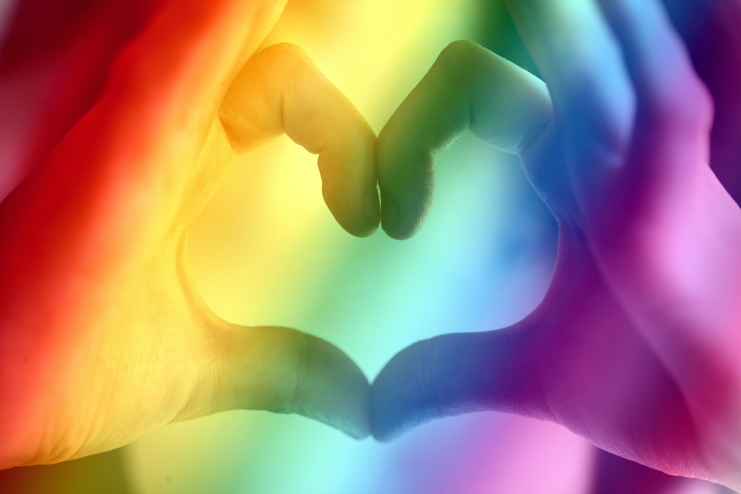 A hand forms a heart behind a translucent rainbow foreground.